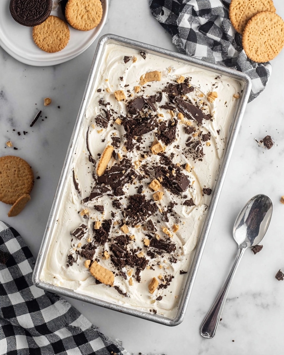 This image shows a rectangular metal container filled with a creamy white dessert that has many pieces of crushed dark chocolate and light-colored cookies spread evenly on top. The dessert surface looks smooth with slight swirls and a mix of dark and light cookie crumbs scattered everywhere giving it a textured look. Around the container, there is a white plate with different types of cookies, a silver spoon on a white marbled surface, and a checkered black and white cloth partially visible. Photo taken with an iphone --ar 4:5 --v 7