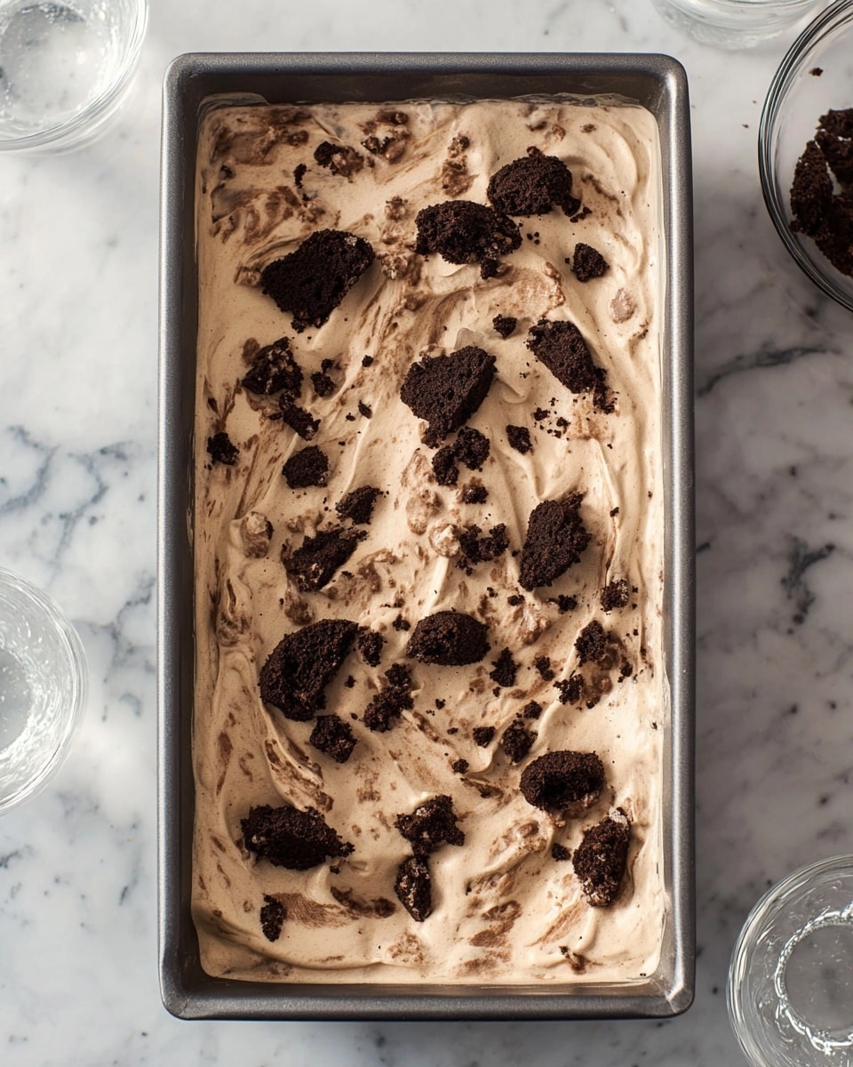 A rectangular gray metal pan filled with one thick layer of light brown creamy dessert that has a smooth, slightly swirled texture. Scattered unevenly on the surface are broken dark brown cookie pieces with a rough and crumbly texture. The pan is placed on a white marbled surface with part of three clear glass bowls visible around the pan’s edges. photo taken with an iphone --ar 4:5 --v 7