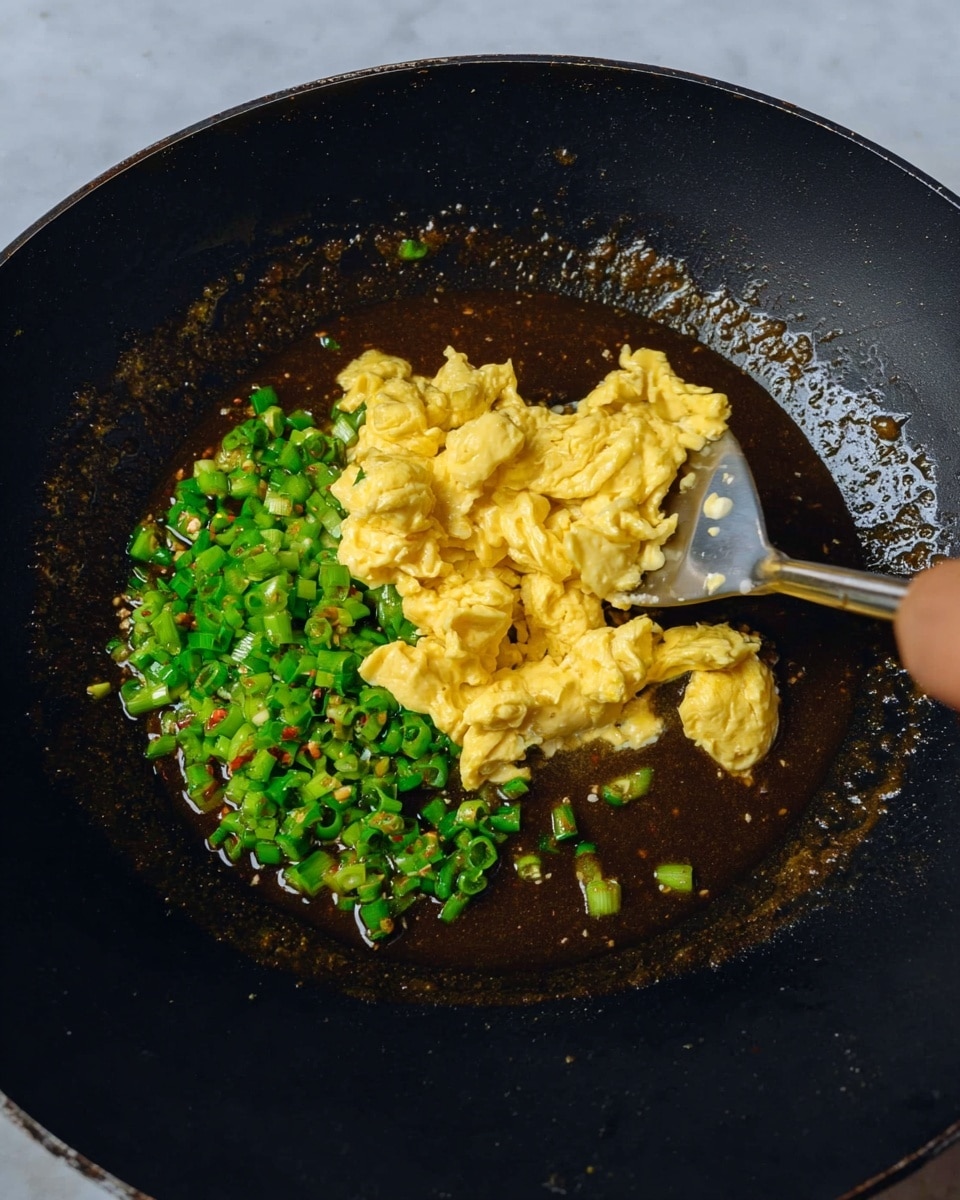 The image shows a cooking pan with a black surface filled with a mixture of food. There are three layers: the bottom layer is a dark brown sauce or liquid that covers the base of the pan; on top of the sauce, to the left side, there is a layer of small, chopped green chili pieces; the top layer, placed roughly in the center, is a pile of cooked yellow scrambled eggs with a soft texture. A silver spatula is lifting some of the scrambled eggs from the pan, with a woman's hand holding the spatula from above. The background around the pan is a white marbled surface. photo taken with an iphone --ar 4:5 --v 7