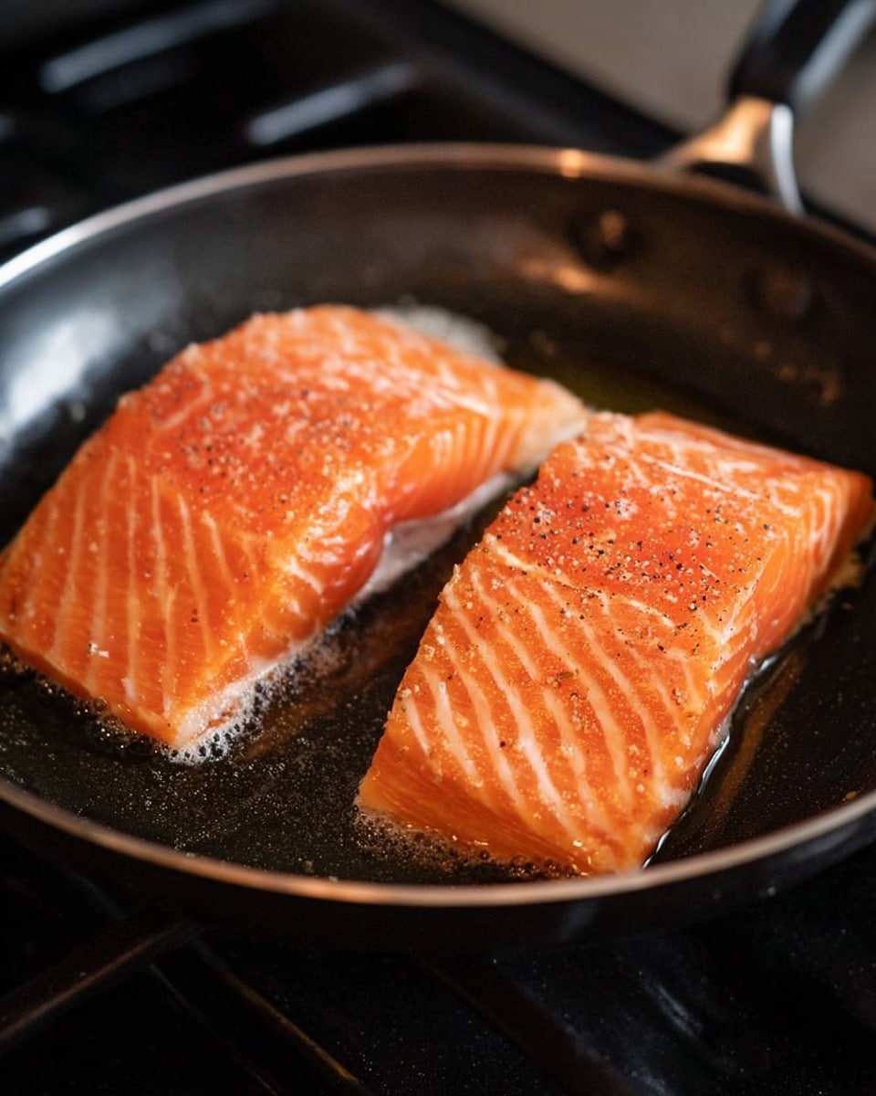 Two thick pieces of raw salmon with bright orange and light white lines lie in a black frying pan with a small amount of oil. The salmon pieces have some coarse salt and black pepper sprinkled on top, adding rough texture to the smooth surface. The frying pan is on a stove burner, and the background shows parts of the stove. The edges of the salmon pieces touch the pan, showing a slight change in color from heat. photo taken with an iphone --ar 4:5 --v 7