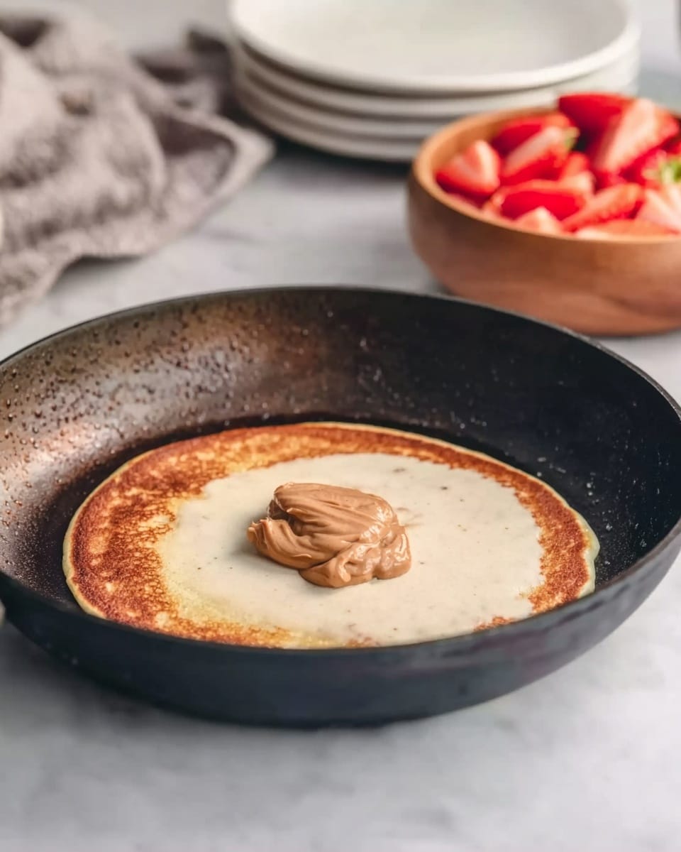 A frying pan with a light golden pancake cooking in the center, topped with a dollop of smooth, light brown spread. The frying pan has a slightly worn edge with a black interior. In the background, there is a wooden bowl filled with sliced strawberries and a stack of white plates on a white marbled surface. A gray cloth is casually placed behind the plates. The scene is lit softly with natural light highlighting the textures. Photo taken with an iphone --ar 4:5 --v 7