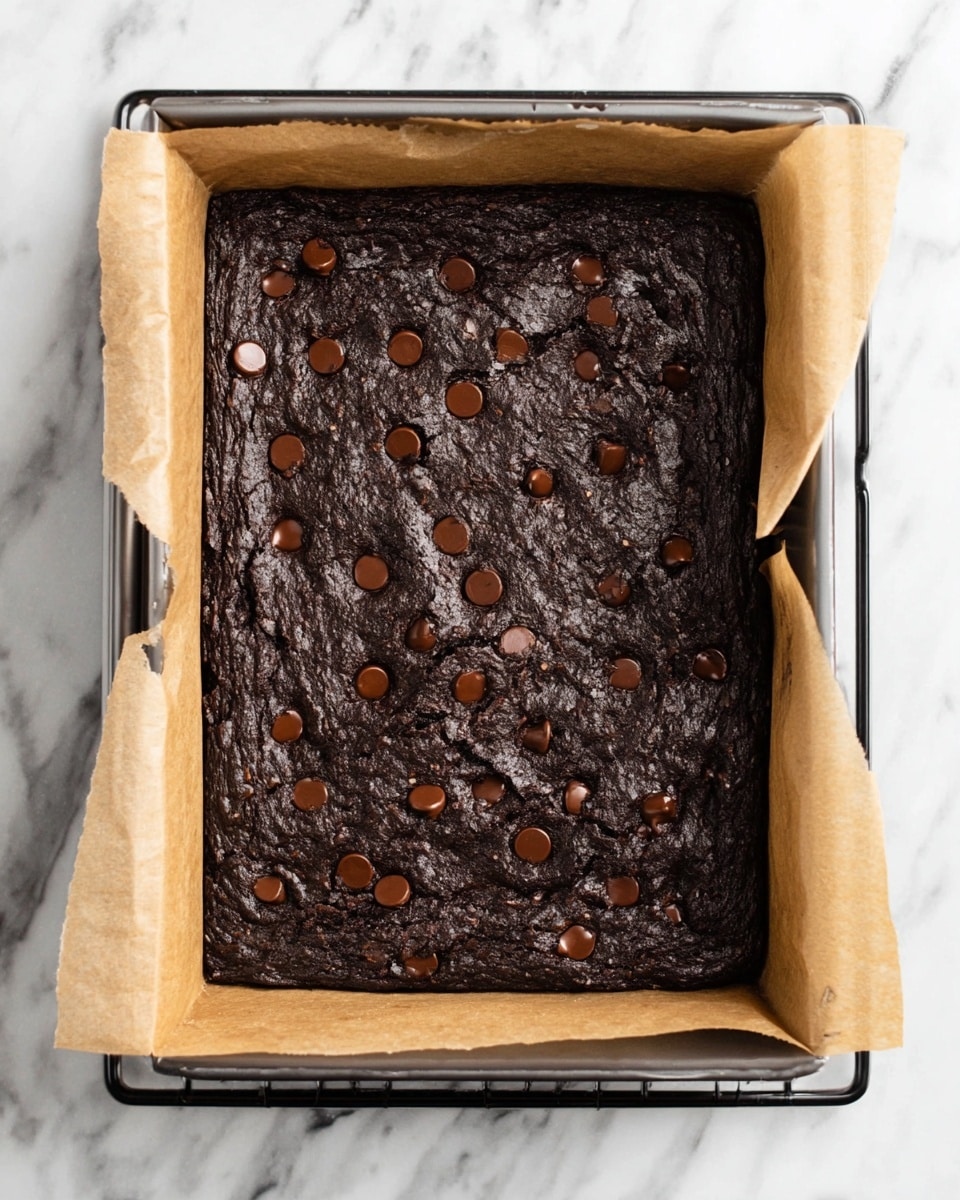 The image shows a rectangular dark brown brownie baked in a metal pan lined with light brown parchment paper that folds over the shorter sides. The brownie has a rough, cracked surface texture with a few glossy chocolate chips partly melted and scattered evenly on top. The pan is placed on a black rack over a white marbled surface. The overall look is rich and dense with a slightly uneven top layer. photo taken with an iphone --ar 4:5 --v 7
