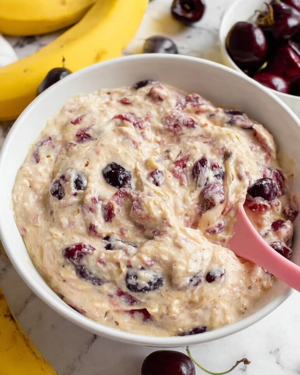 A white bowl filled with a thick, creamy mixture that has a light beige color with dark purple and red bits scattered throughout, showing chunks of fruit or berries mixed inside. A pink spoon is partially dipped into the mixture, resting on the side of the bowl. In the background, there are some yellow bananas and a white bowl with dark red cherries. The whole scene is set on a white marbled surface. photo taken with an iphone --ar 4:5 --v 7