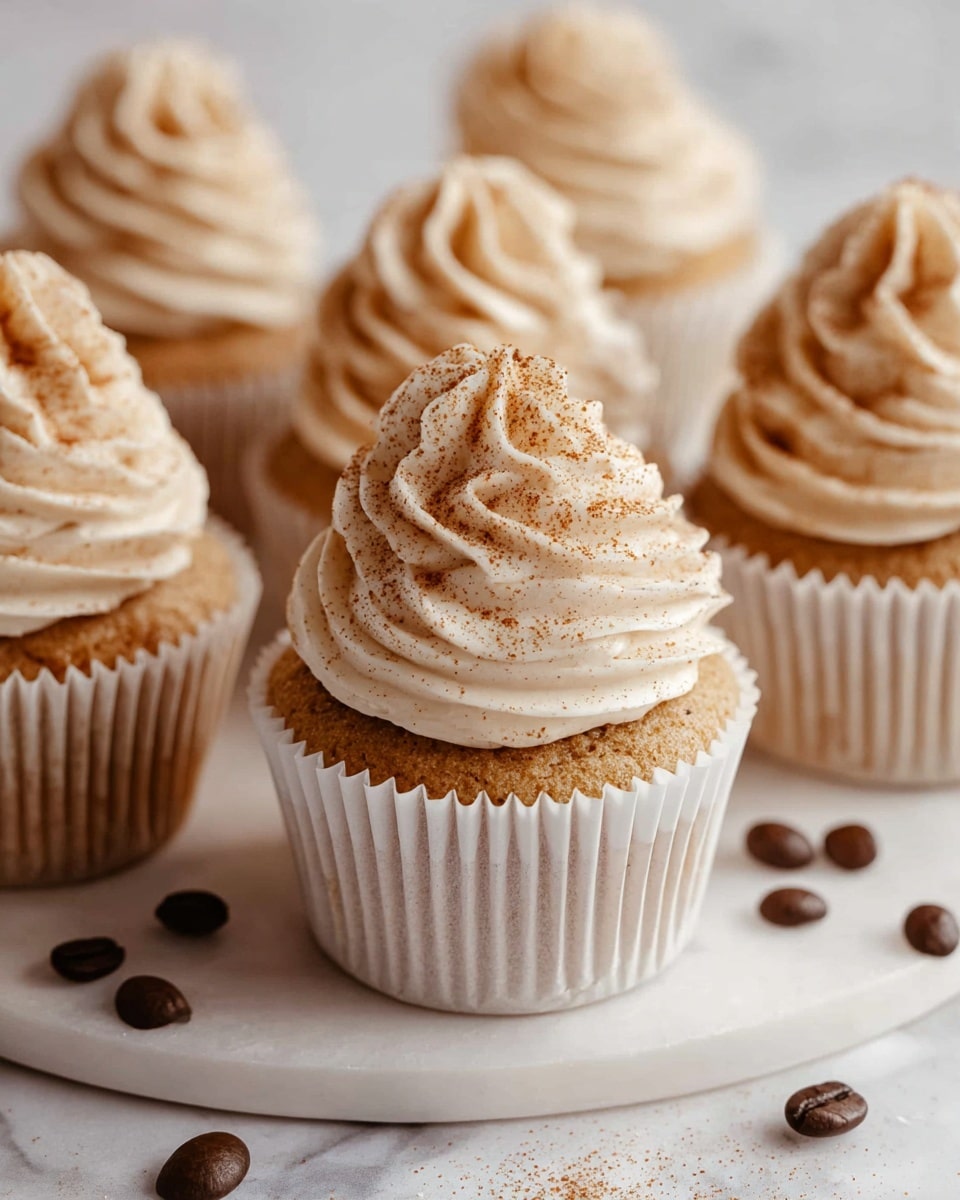 There are several cupcakes arranged closely, each with a light brown base wrapped in white paper liners. On top of each cupcake is a tall swirl of creamy beige frosting with a soft texture, sprinkled lightly with brown powder, giving a cinnamon or coffee-like appearance. The cupcakes sit on a round white plate with coffee beans scattered around it. The surface beneath the plate has a white marbled texture, adding a clean and elegant look to the scene. photo taken with an iphone --ar 4:5 --v 7
