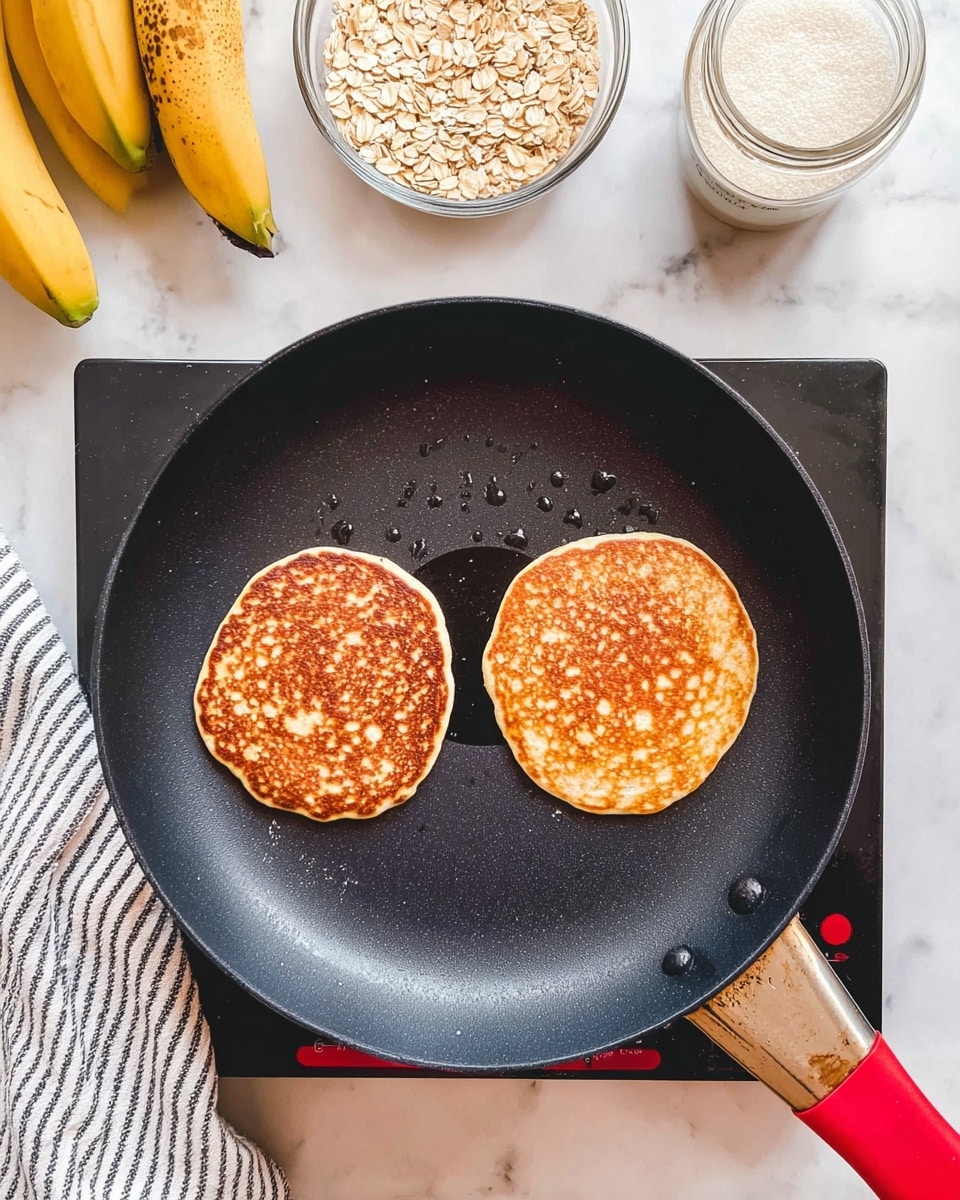 Two golden-brown pancakes with slightly crispy edges cook side by side in a black nonstick frying pan with a red circle near the center, sitting on a black stovetop. The pancakes have a warm, toasted texture with small air bubbles on top. Around the pan, there are bananas with brown spots at the bottom left, a glass bowl filled with oats at the bottom center, and a glass jar with a white powdery substance near the top. A striped cloth is partially visible on the left side, all set on a white marbled surface. Photo taken with an iphone --ar 4:5 --v 7