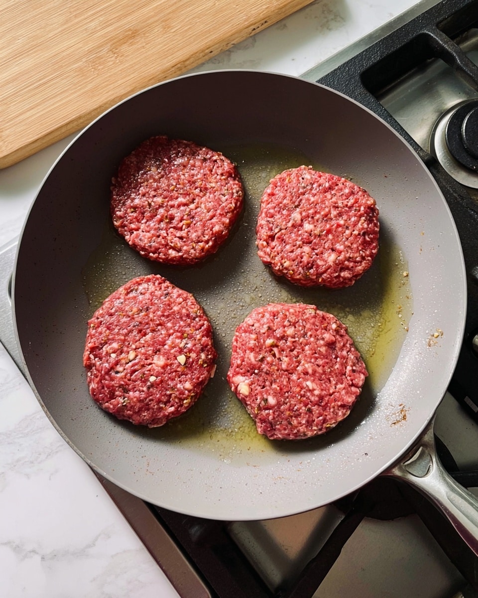Four thick raw burger patties with visible bits of seasoning and small white pieces are arranged evenly inside a large gray non-stick pan. Each patty has a rough textured surface and sits slightly on top of a small pool of oil. The pan rests on a metal stove with black knobs and a silver burner grill, next to a light wooden cutting board on a white marbled surface. Photo taken with an iphone --ar 4:5 --v 7