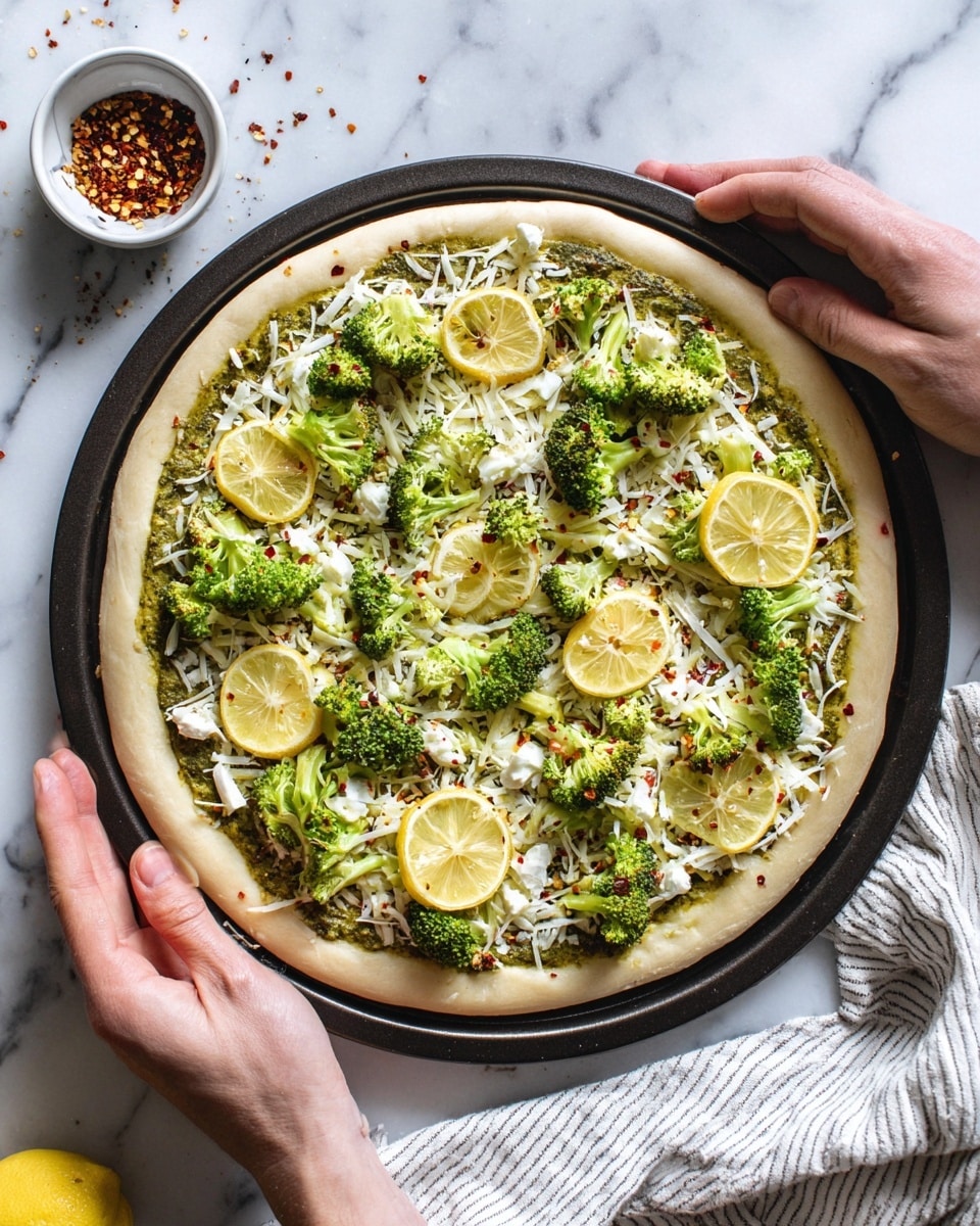 A round pizza with a thick light beige crust sits on a black pizza pan, held by two hands at the edges. The pizza has several layers: the bottom layer is a green pesto spread, topped with a thin layer of white shredded cheese evenly scattered. On top, there are small white dollops of soft cheese, bright green broccoli florets spread around, and thin yellow lemon slices evenly placed. Small red chili flakes are sprinkled all over the pizza. The setting is on a white marbled surface with a white and gray striped cloth on the bottom right corner, and a small white bowl with chili flakes is visible on the bottom left. Photo taken with an iphone --ar 4:5 --v 7