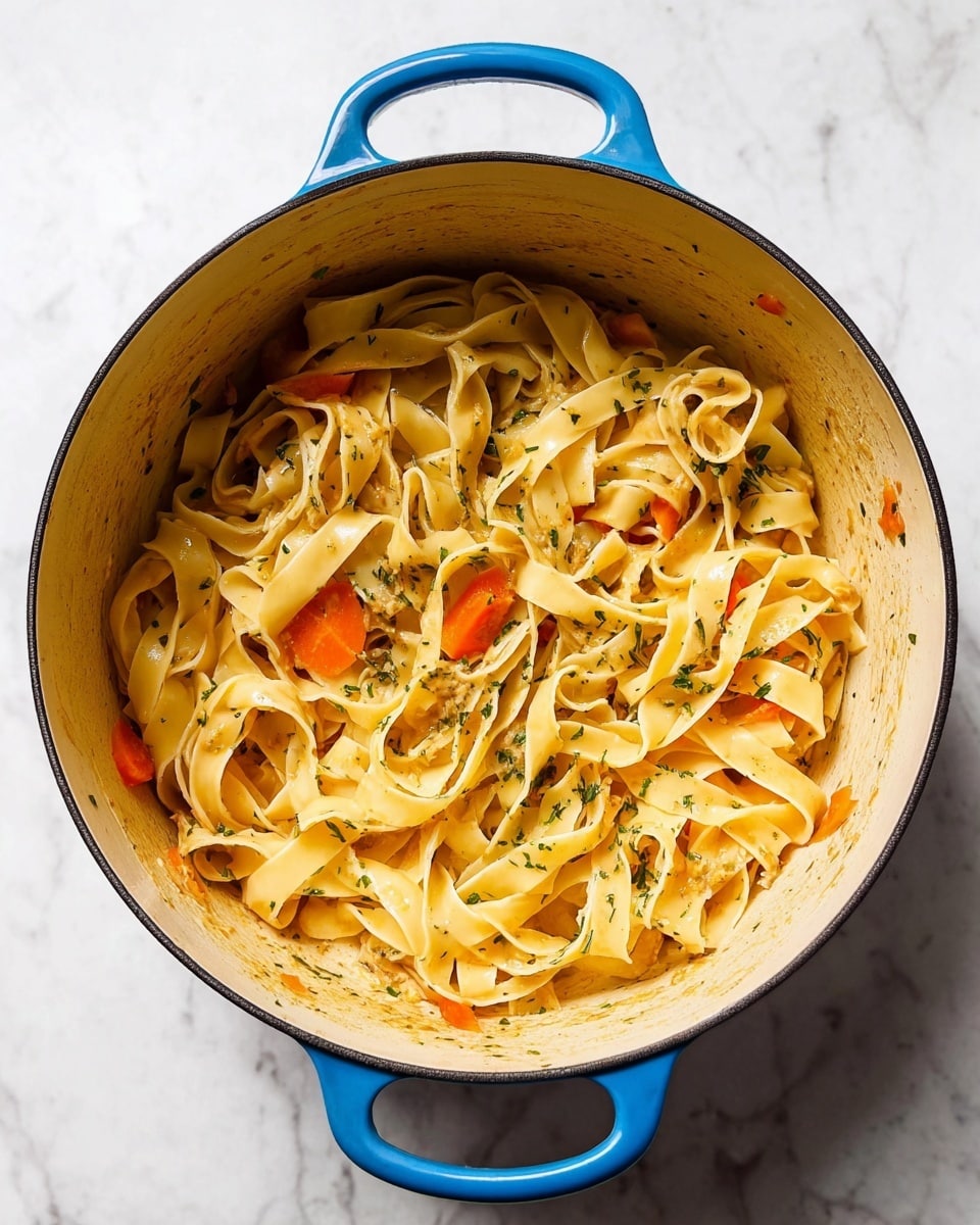 The image shows a blue pot filled with wide, flat fettuccine pasta mixed with small slices of orange carrots and sprinkled with green herbs. The pasta looks soft and slippery, with each ribbon curled and layered on top of one another, giving a light yellow color with a shine from the sauce. The carrot bits add a bright pop of orange, scattered throughout the pot, while the green herbs are lightly sprinkled on top acting as a fine garnish. The inside of the pot has some sauce stuck to the sides, showing the mixing process. The pot sits on a white marbled surface, creating a clean and fresh background. photo taken with an iphone --ar 4:5 --v 7