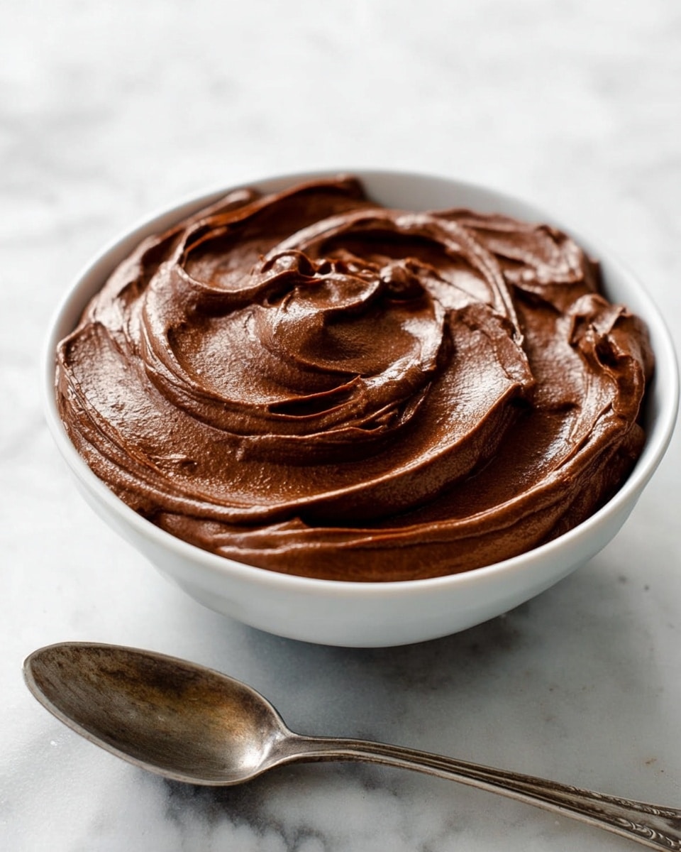 A white bowl filled with a smooth, thick layer of rich dark brown chocolate mousse, swirled with soft peaks and glossy waves that create a textured, creamy surface. The bowl sits on a white marbled surface, and next to it is an old-fashioned silver spoon stained slightly with the same chocolate mousse. The mousse looks light and airy, with whipped curves catching soft light, showing its velvety texture and depth. photo taken with an iphone --ar 4:5 --v 7