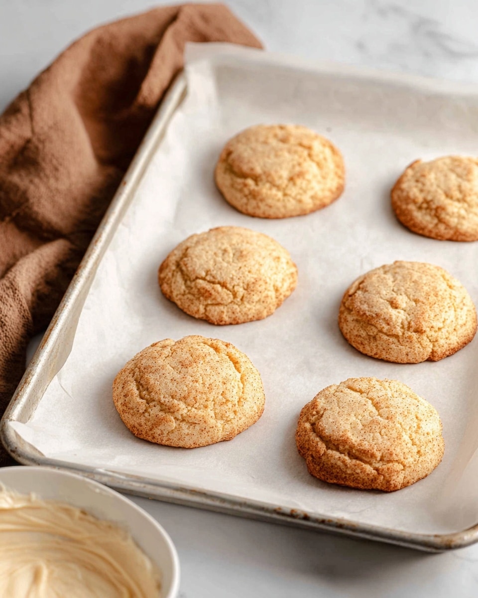 Six round, light brown cookies with a slightly cracked and rough texture are spaced evenly on a sheet of white parchment paper that lines a silver baking tray. The tray rests on a white marbled surface, with a soft brown cloth partially visible in the top left corner and a white bowl with creamy batter in the bottom left corner of the image. The cookies have a soft, slightly raised middle and a rougher edge. Photo taken with an iphone --ar 4:5 --v 7