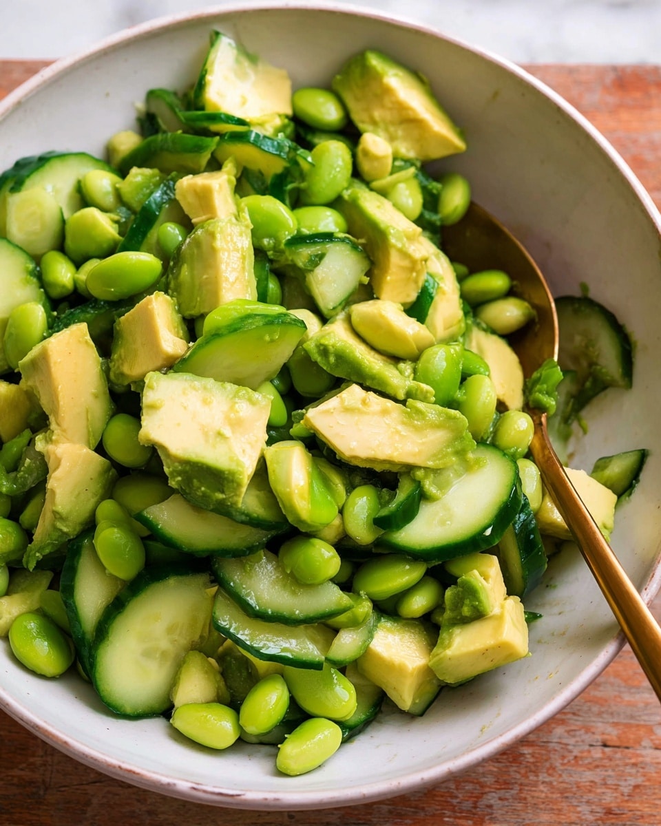 A close-up of a white bowl filled with a fresh salad showing three main layers: chunky avocado pieces in light green and yellow shades with a soft texture, bright green edamame beans that look smooth and round, and chopped cucumber slices with dark green skin and pale green inside, all mixed together. A gold spoon rests inside the bowl, partially covered by the salad. The white bowl sits on a wooden surface, but the background is changed to a white marbled texture photo taken with an iphone --ar 4:5 --v 7