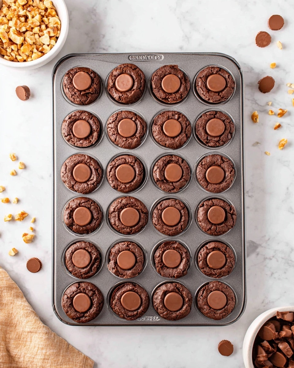 A metal baking tray filled with 24 small, round chocolate brownies, each with a smooth, circular chocolate piece pressed into the center on top. The brownies have a cracked, slightly rough texture on top and a rich dark brown color. They are arranged in a 6 by 4 grid inside the tray. The tray sits on a white marbled surface with some small chocolate and nut pieces scattered nearby. A white bowl filled with chopped nuts is visible at the bottom left, and another white bowl with round chocolate pieces is at the bottom right. Photo taken with an iphone --ar 4:5 --v 7