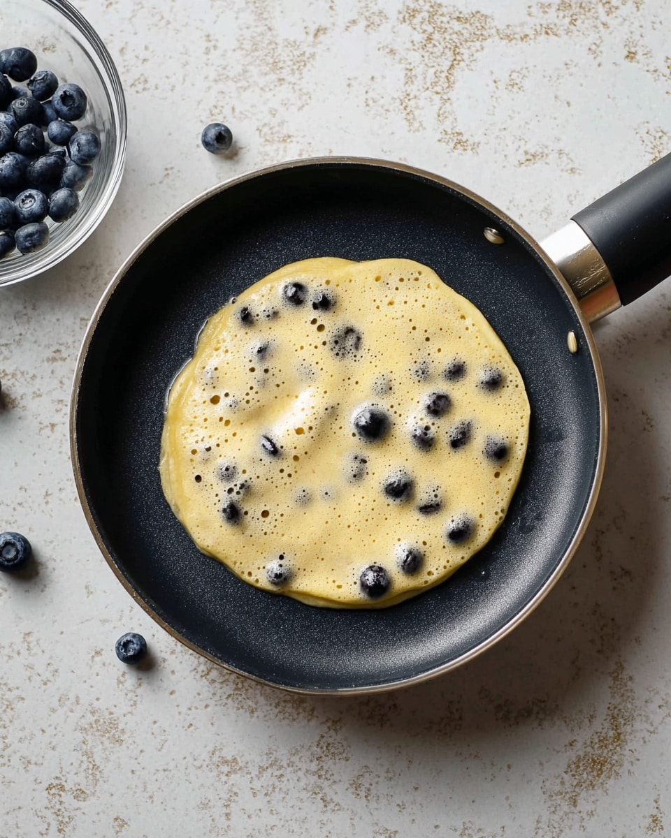A small, round pancake batter with blueberries inside is cooking on a black pan. The batter is light yellow with small holes and visible blueberries scattered throughout, some partly submerged and some on the surface. Next to the pan on the white marbled texture surface is a clear bowl with more of the same batter and blueberries. Photo taken with an iphone --ar 4:5 --v 7
