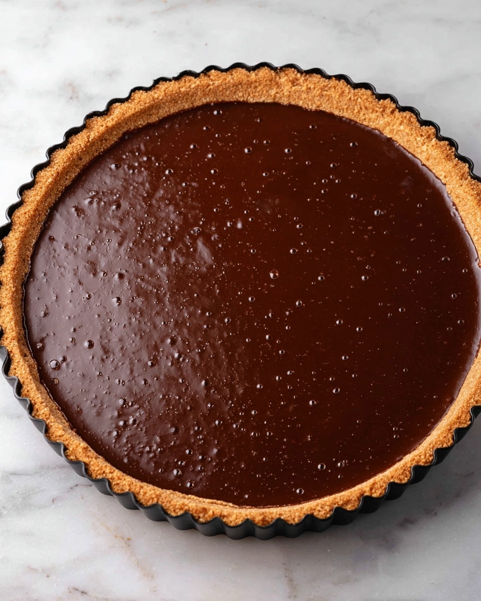 A round pie with two layers is shown on a white marbled surface. The bottom layer is a light brown crust with a crumbly texture, forming a thick edge around the pie that is slightly scalloped. The top layer is a smooth, dark brown chocolate filling, shiny and even, filling the crust completely with tiny bubbles visible on the surface. The pie is still in a black tin with scalloped edges just outside the crust photo taken with an iphone --ar 4:5 --v 7