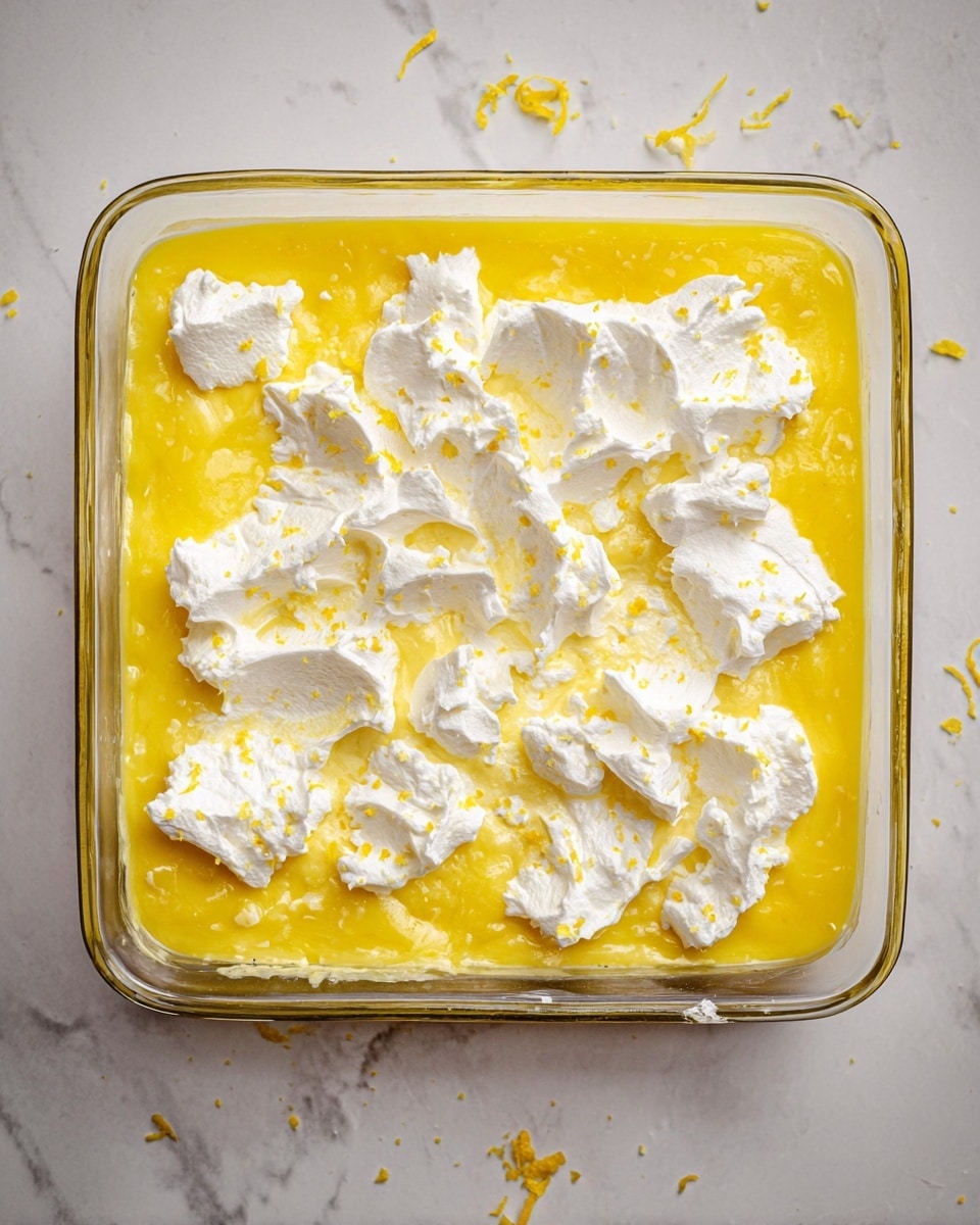 A clear glass square baking dish filled with a bright yellow smooth lemon layer as the base, topped with scattered dollops of white fluffy cream topping spread unevenly across the surface. The cream has a soft texture with slight peaks and tiny specks of yellow lemon zest sprinkled over it. The dish rests on a white marbled surface with small bits of lemon zest around it, showing a clean and fresh look. photo taken with an iphone --ar 4:5 --v 7