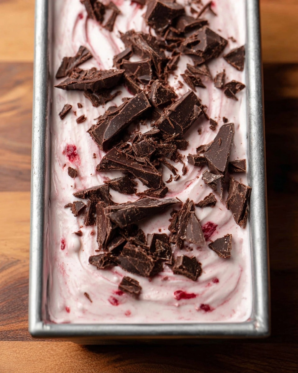 The image shows a close-up of a dessert in a rectangular metal pan, with one thick layer of light pink creamy mixture swirled with bits of red inside, topped with large, rough pieces of dark brown chocolate sprinkled unevenly across the surface. The creamy layer looks smooth and soft, while the chocolate pieces add a textured contrast. The pan is on a wooden surface, but the background is changed to a white marbled texture. photo taken with an iphone --ar 4:5 --v 7