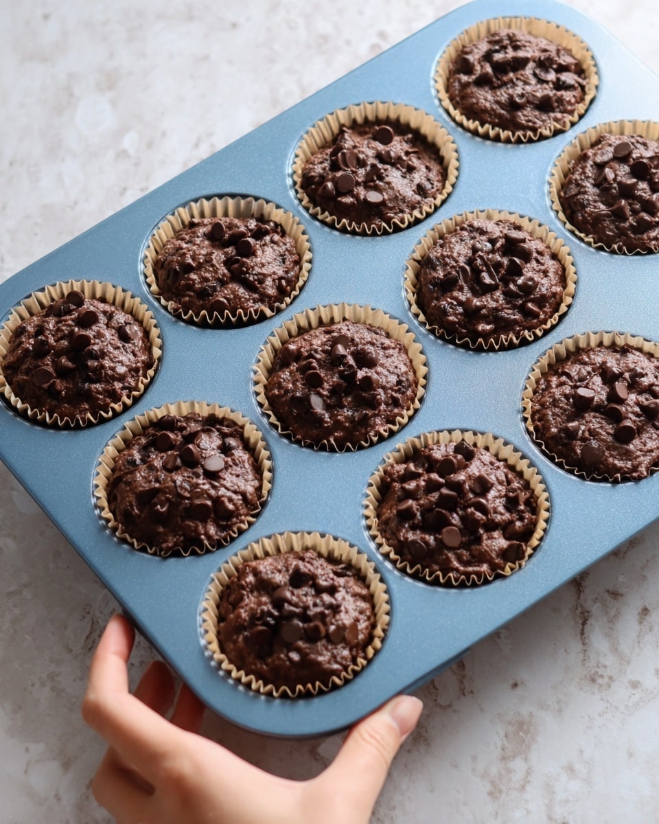 A close-up view of a blue muffin tray filled with nine unbaked chocolate muffins in light brown paper liners. Each muffin has a thick, dark brown chocolate batter with visible chocolate chips scattered on top. The surface of the batter is bumpy and moist-looking. A woman's hand is holding the corner of the tray from the bottom left. The tray sits on a white marbled surface. photo taken with an iphone --ar 4:5 --v 7