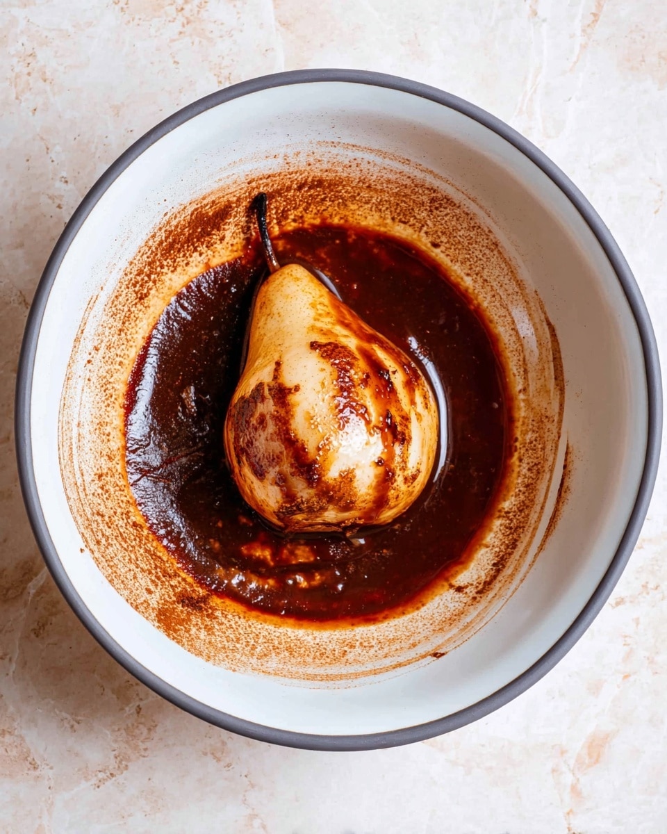 A white bowl with a dark gray rim holds one cooked pear placed in the center. The pear has a light brown skin with darker, slightly charred spots and is partly covered with a thick dark brown sauce that spreads over the bottom of the bowl, leaving brown streaks along the sides. The surrounding sauce is rich and glossy, with a powdery texture visible on the edges. The bowl sits on a white marbled surface. photo taken with an iphone --ar 4:5 --v 7
