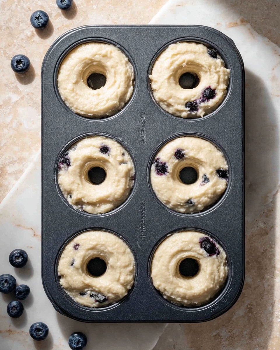 A dark gray donut baking tray holds six rings of pale, creamy batter, each ring showing soft, uneven textures with small dark spots of blueberries scattered inside. The rings are well-shaped with a visible hole in the center. The tray rests on a white marbled surface, with a few fresh blueberries placed casually nearby. Soft shadows fall across the scene, adding subtle depth. photo taken with an iphone --ar 4:5 --v 7