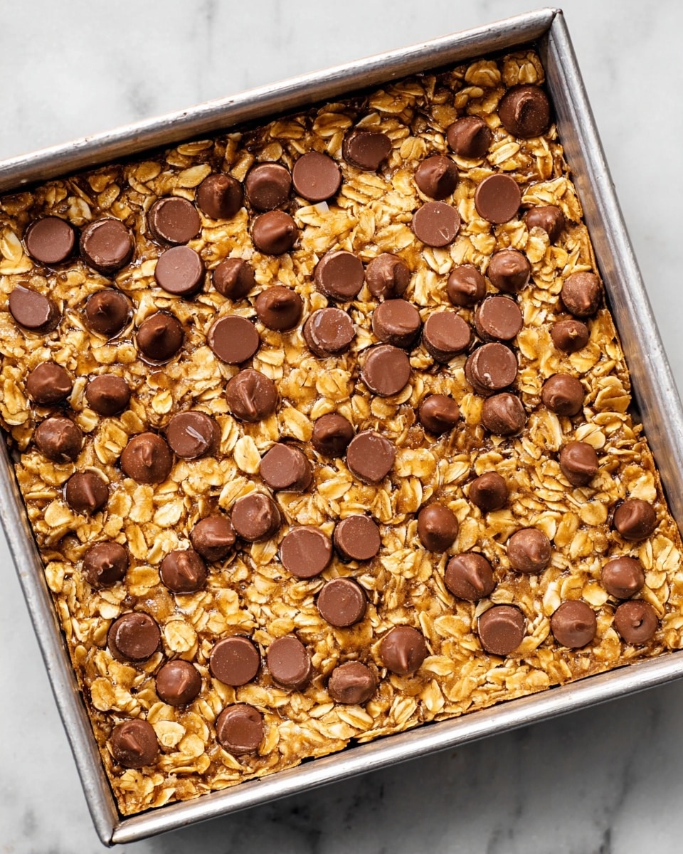 The image shows a single-layer oat bar in a square metal pan. The base is made up of light golden oats that have a slightly glossy texture, as if mixed with a syrup or melted butter. Scattered evenly across the top are many glossy dark brown chocolate chips, some sitting upright while others are slightly melted and flat. The edges of the oat bar are a bit darker, showing slight crispiness. The background is a white marbled surface. photo taken with an iphone --ar 4:5 --v 7