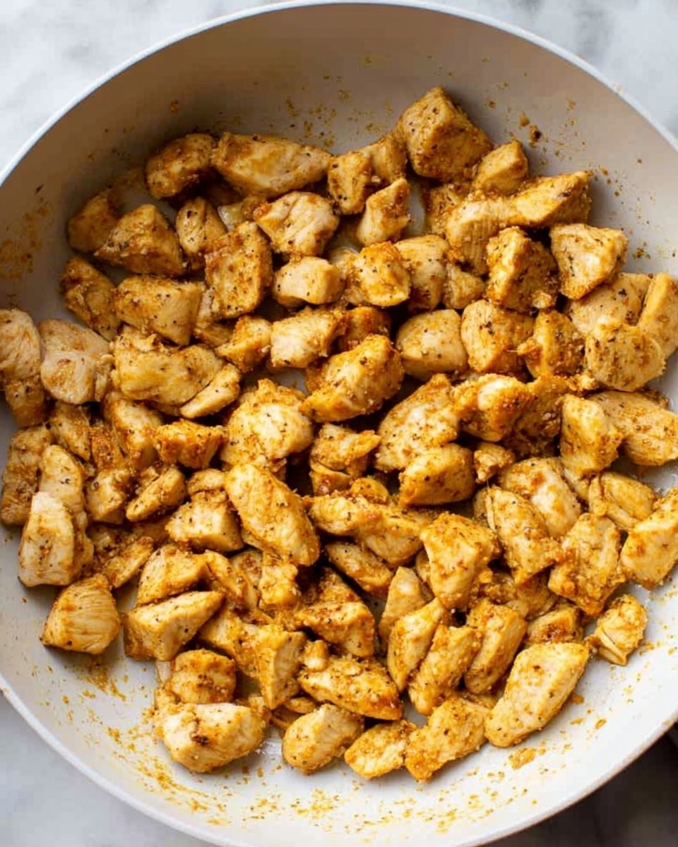 The image shows a white pan filled with small, cooked pieces of chicken seasoned with spices. Each piece is golden brown with a slightly crispy texture on the outside. The chicken pieces are spread evenly across the pan, with some small bits of seasoning visible on the pan's surface. The background is a white marbled texture. Photo taken with an iphone --ar 4:5 --v 7