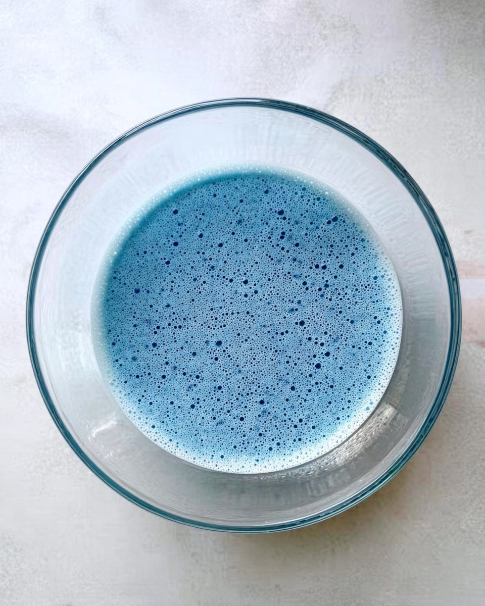 A clear glass bowl filled with a single layer of light blue liquid that has a frothy, bubbly texture on the surface, small bubbles distributed evenly across it, sitting on a white marbled surface background. photo taken with an iphone --ar 4:5 --v 7