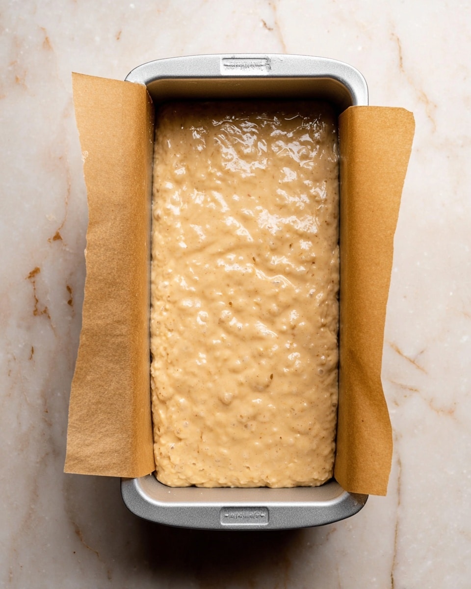 A rectangular silver baking pan lined with light brown parchment paper holds a single layer of thick, light brown batter with small bubbles and a slightly uneven texture on the surface. The pan rests on a white marbled surface. The batter fills the pan almost to the top edges, showing a creamy, slightly lumpy consistency. photo taken with an iphone --ar 4:5 --v 7