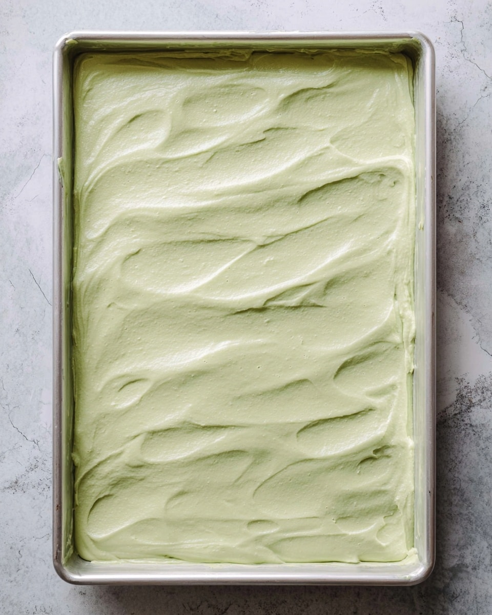 A single flat layer of smooth, pale green creamy mixture spread evenly across a large silver baking tray, with soft light texture and slight undulating waves on the surface. The tray sits on a white marbled textured background. photo taken with an iphone --ar 4:5 --v 7