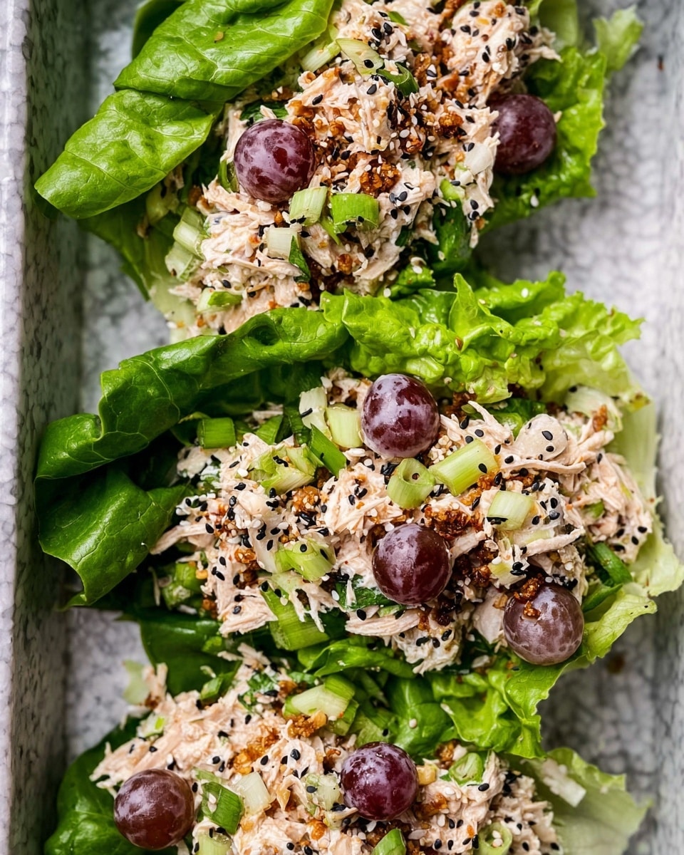 The image shows three lettuce wraps placed close to each other inside a tray with a white marbled texture background. Each wrap has one thick layer of bright green lettuce leaves at the bottom, topped with a creamy mixture that is off-white with textures of shredded meat or vegetable, light purple grape halves, and small green slices that look like chopped spring onions. The creamy mixture is sprinkled with black and white sesame seeds and bits of a toasted topping, giving it a contrasting texture and colors. The wraps are neat, colorful, and visually fresh. photo taken with an iphone --ar 4:5 --v 7