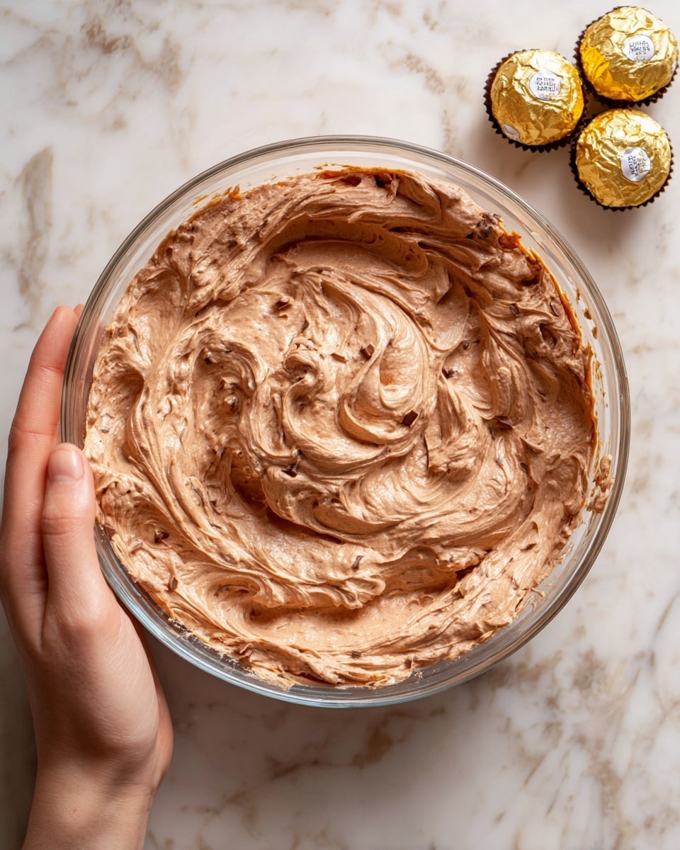 A top-down view shows a clear glass bowl filled with a thick, light brown chocolate mixture with swirled texture and small dark spots around the bowl edges, placed on a white marbled surface with a woman's hand holding the bowl on the left. Above the bowl, two round chocolate candies in gold wrappers sit side by side. The background reveals some nested white bowls in the top left corner and a wooden item with visible grains in the top right corner. photo taken with an iphone --ar 4:5 --v 7