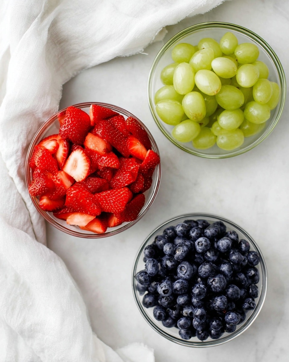 Three clear glass bowls sit on a white marbled surface, each filled with different fruits. The top right bowl holds smooth, light green grapes with a shiny texture. The center bowl is full of bright red strawberries, sliced into small pieces showing their juicy inside. The bottom right bowl contains dark blue blueberries with a slightly matte finish. A soft, white cloth is casually placed to the left of the bowls, adding a gentle texture to the scene. photo taken with an iphone --ar 4:5 --v 7