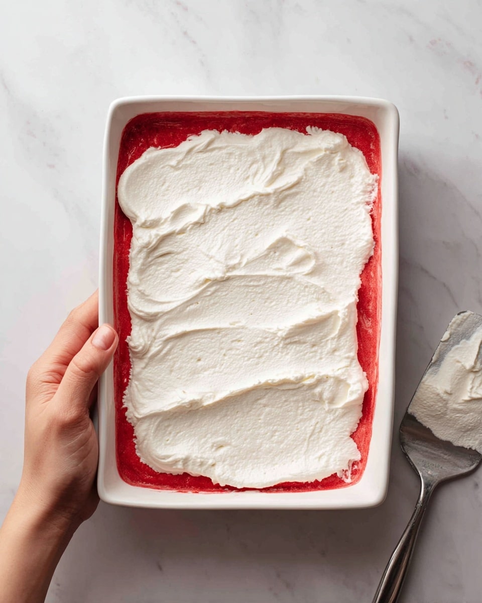 A white rectangular dish sits on a white marbled surface, filled with one thick red layer at the bottom and a thick white cream layer spread unevenly on top, with soft peaks and textured ridges. A woman's hand holds the dish carefully from the top right corner. A spatula with some white cream rests to the bottom right of the dish. photo taken with an iphone --ar 4:5 --v 7