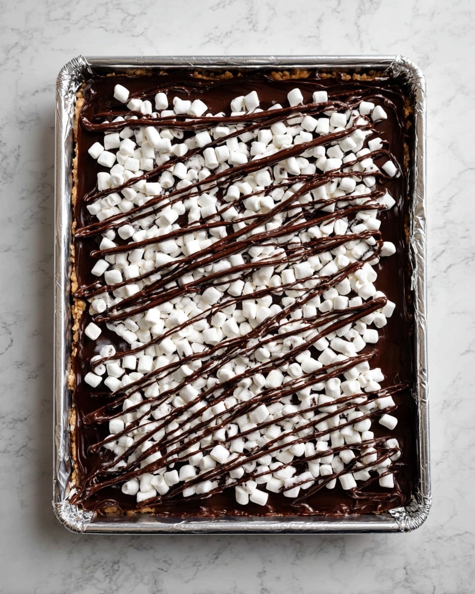 A rectangular baking tray lined with foil holds a layered dessert. The bottom layer is lightly visible nuts or base, topped with a thick layer of dark, smooth chocolate spread. On top, there is a scattered layer of small, white mini marshmallows covering the chocolate. Over the marshmallows, thin, dark chocolate is drizzled in random, overlapping lines creating a web-like pattern. The tray sits on a surface with a white marbled texture photo taken with an iphone --ar 4:5 --v 7