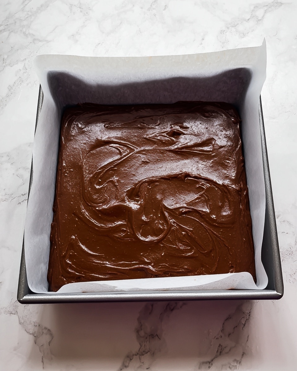 A square metal baking pan lined with white parchment paper holds a thick, smooth layer of shiny dark brown chocolate batter spread evenly across the surface. The batter has slight swirls and small lumps, giving it a rich, creamy texture. The pan is placed on a white marbled surface with soft gray veining. photo taken with an iphone --ar 4:5 --v 7
