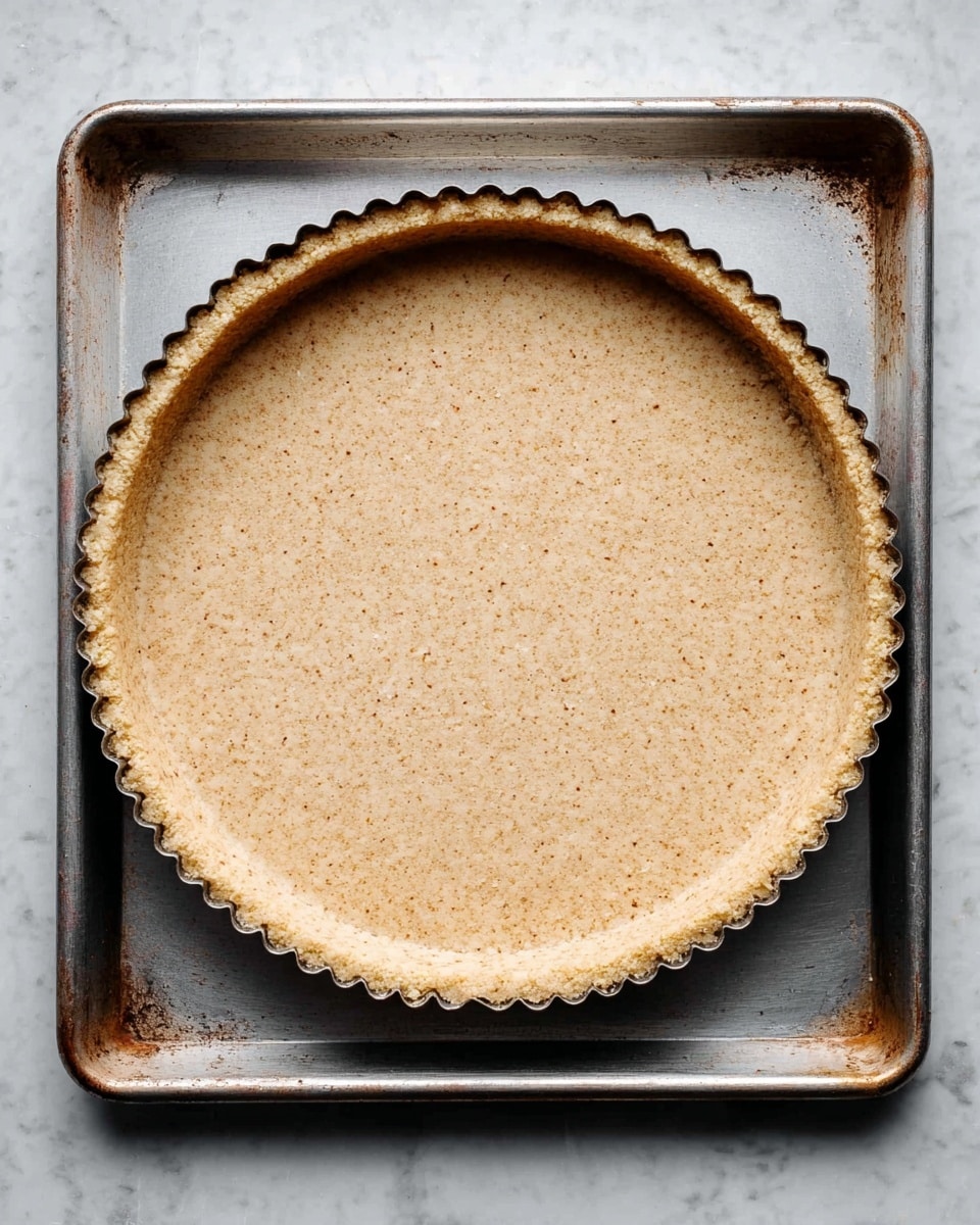 A single round tart crust with a fluted edge sits in a tart pan, displaying an even, light beige color with small brown specks throughout its grainy texture. The crust layer is smooth and pressed firmly against the pan's sides, resting on a slightly distressed metal baking tray. The entire scene is set on a white marbled surface. Photo taken with an iphone --ar 4:5 --v 7
