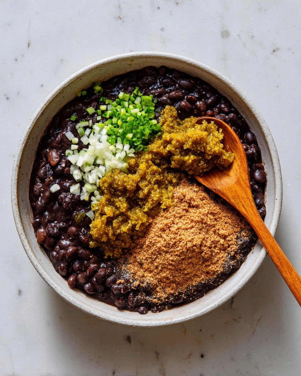 A bowl filled with cooked black beans forming a dark, glossy base layer. On top, there are three distinct toppings spread in separate sections: finely chopped white and green onions with a fresh texture, a golden-yellow chunky relish with a slightly wet appearance, and a mound of light brown finely ground spice or powder. A wooden spoon with a smooth light brown handle and flat end rests inside the bowl on the right side. The bowl is white and placed on a white marbled surface. Photo taken with an iphone --ar 4:5 --v 7