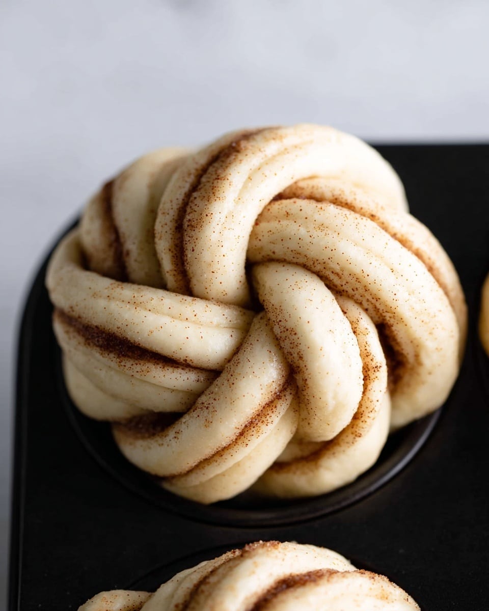 The image shows a close-up of a knotted dough bun with three main twisted layers, each layer pale beige with a soft, smooth texture. Between these layers, there are thin streaks of brown cinnamon sugar filling visible throughout the knot, giving a speckled effect of spices. The bun sits snugly in a black baking tray cup with a white marbled background blurred above. Photo taken with an iphone --ar 4:5 --v 7