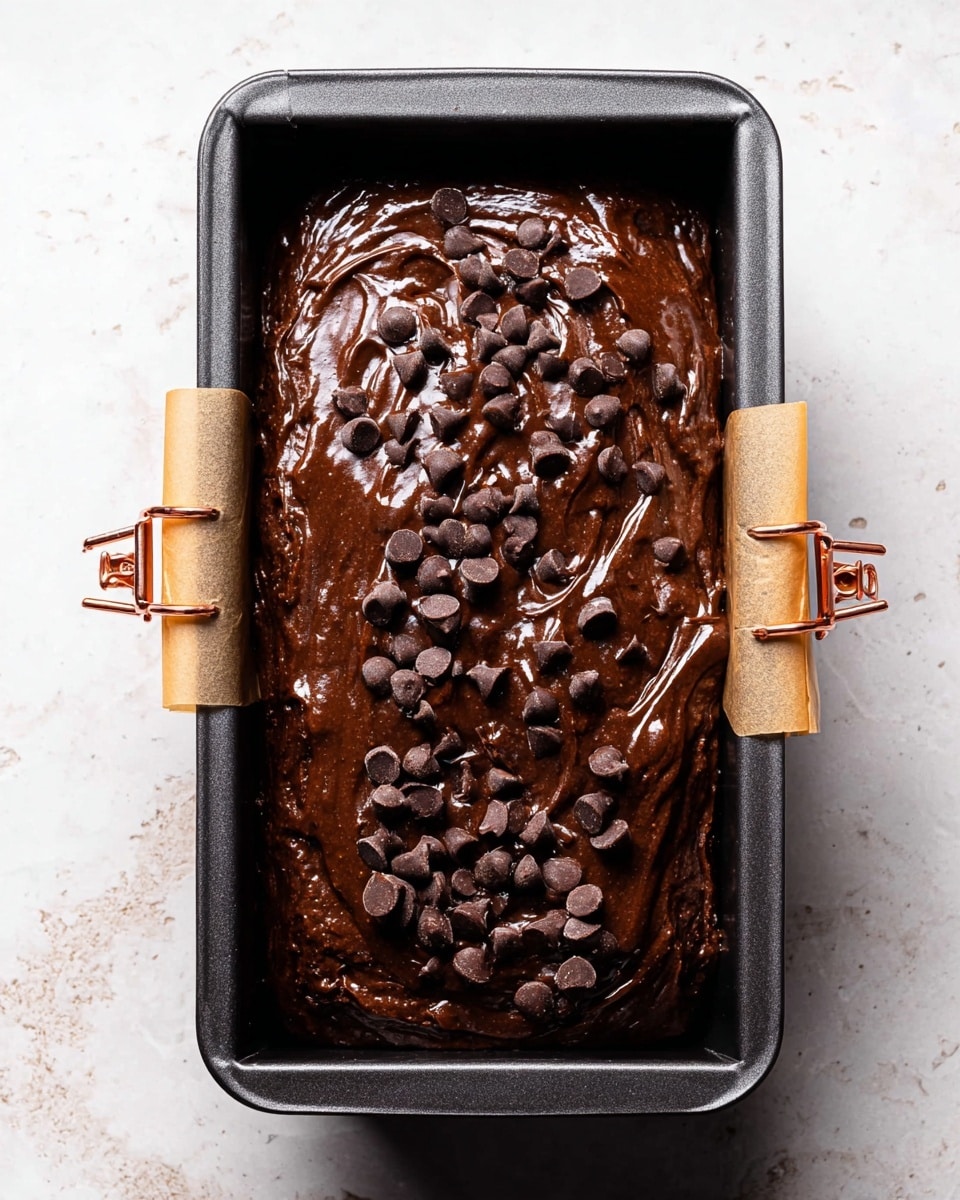 A dark brown chocolate batter fills a rectangular black metal loaf pan lined with parchment paper that folds over the short sides, held with two copper clips. The batter is thick and unevenly spread, with small ridges and waves, topped with scattered milk and dark chocolate chips that contrast with the shiny, wet surface. The pan rests on a surface with a white marbled texture, adding a subtle light background to the rich chocolate scene. photo taken with an iphone --ar 4:5 --v 7