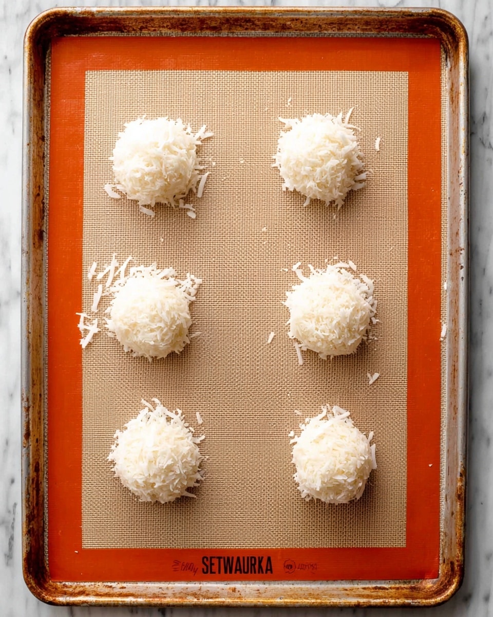 Six small, round mounds of white shredded coconut are placed evenly in two rows of three on a silicone baking mat with orange borders. The mat is set on a metal baking sheet with a worn look, and the background beneath the sheet is a white marbled texture. Each coconut mound looks soft and slightly fluffy, with small, fine strands sticking out around the edges, giving a textured and delicate appearance. Photo taken with an iphone --ar 4:5 --v 7