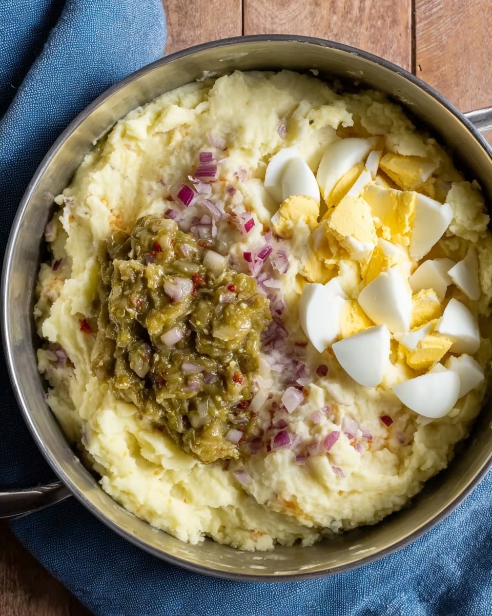 A close-up view of a round metal bowl filled with creamy mashed potatoes mixed with small red onion pieces covering the bottom layer. On the top right side is a pile of chopped hard-boiled eggs showing white and yellow colors, while on the top left side sits a chunky greenish relish with bits of red visible inside. The bowl is placed on a blue cloth on a wooden surface, with the creamy texture of the mashed potatoes clearly visible around the edges. photo taken with an iphone --ar 4:5 --v 7