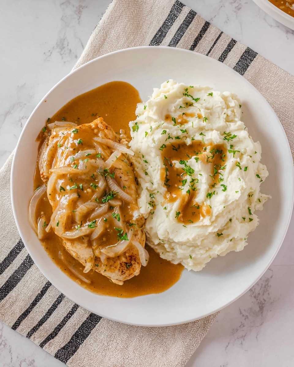 A white plate on a white marbled surface holds a simple meal with two main parts. On the left side, there is a piece of cooked chicken covered in brown gravy mixed with slices of cooked onions and chopped green herbs scattered on top. On the right side, there is a generous mound of white mashed potatoes with smooth and creamy texture, topped with a drizzle of the same brown onion gravy and a few green herbs for garnish. The plate sits on a beige cloth with black stripes under the white marbled surface. Photo taken with an iphone --ar 4:5 --v 7