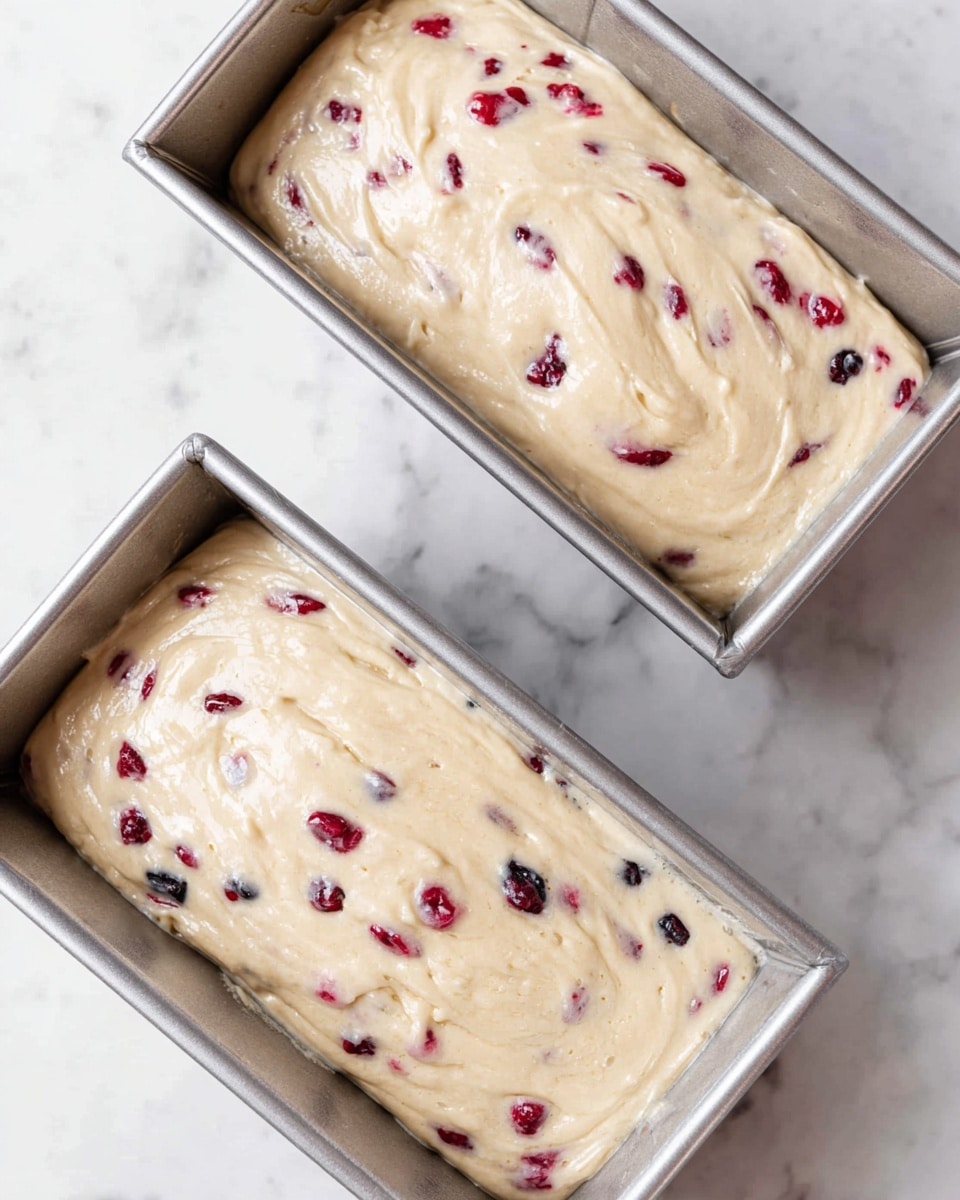 Two rectangular silver baking pans sit on a white marbled surface. Each pan is filled with a creamy, light beige batter that has a smooth, slightly swirled texture on top. Small red and dark purple berry pieces are scattered evenly throughout the batter, creating bright spots of color within the pale mixture. The pans are placed close together, with one slightly above and angled toward the other. photo taken with an iphone --ar 4:5 --v 7