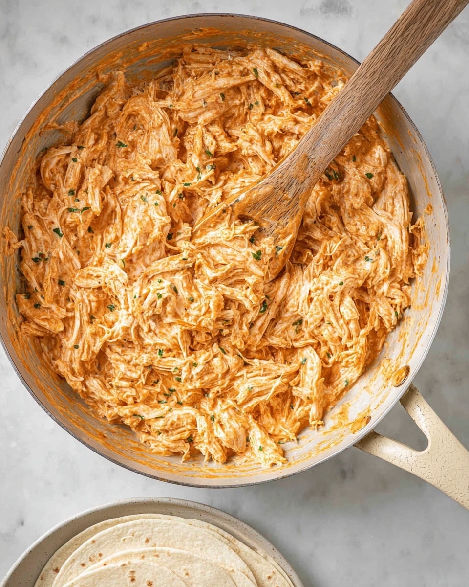 The image shows a close-up of shredded chicken mixed with a creamy orange sauce in a large metal pan with a beige handle. The mixture has a textured look with small green herb bits spread throughout, giving a fresh contrast. A wooden spoon, partially covered with the sauce, rests inside the pan on top of the shredded chicken. Below the pan, a stack of light beige tortillas sits on a white plate, all set on a white marbled surface. photo taken with an iphone --ar 4:5 --v 7