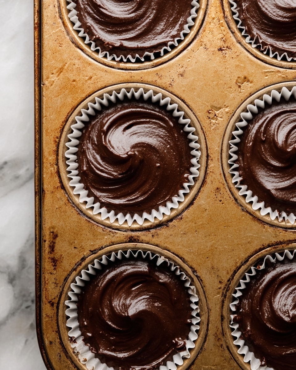 A close-up image shows a metal cupcake tray with six cups filled with thick, dark chocolate batter. Each cup is lined with a white paper liner that has ridged edges, holding the smooth, shiny batter that swirls gently on top. The metal tray has a worn, golden tone with some brown spots and scratches, showing signs of use. The background around the tray is a white marbled texture. photo taken with an iphone --ar 4:5 --v 7