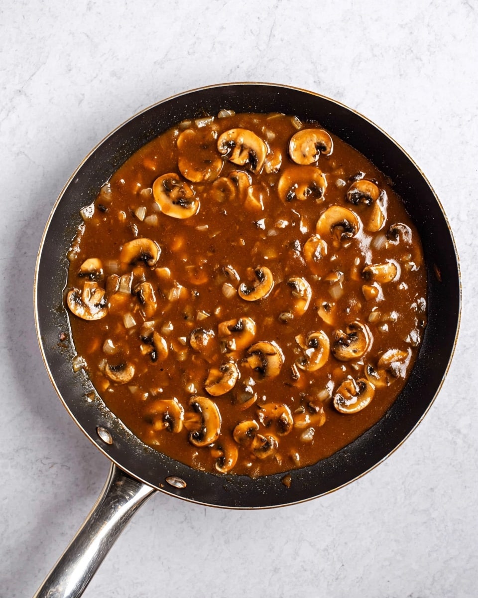A black frying pan with a silver handle is filled with a thick brown mushroom sauce. The sauce has many sliced mushrooms that are light brown with dark centers, floating evenly in the glossy sauce. Small pieces of onion are mixed throughout the sauce, adding texture. The pan sits on a white marbled surface, and the sauce fills the pan nearly to the edges, showing some slight bubbles on top. photo taken with an iphone --ar 4:5 --v 7