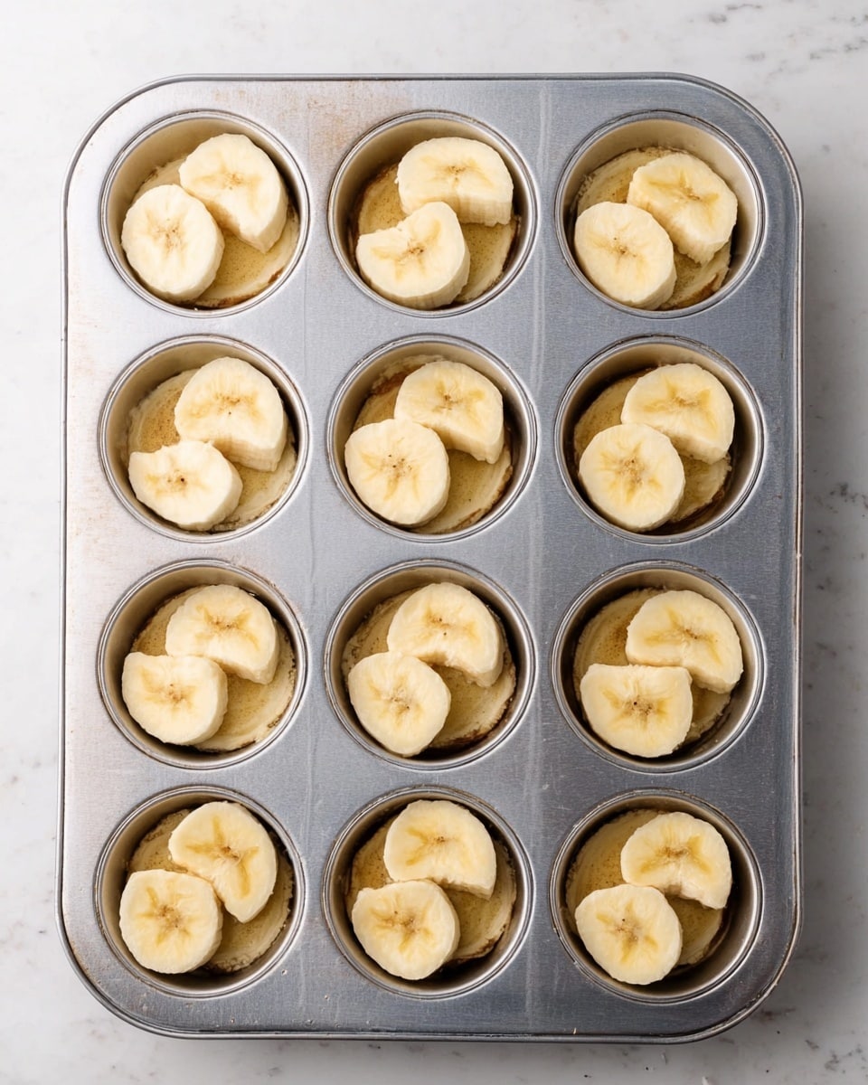 A metal muffin tray holds twelve round cavities, each lined with three banana slices. In each cavity, two banana slices are placed flat at the bottom, with a third slice curved along the side, forming a semi-circle around the other two. The bananas are pale yellow with some light brown spots, showing ripeness. The muffin tray sits on a white marbled surface, and the image is bright and clear. photo taken with an iphone --ar 4:5 --v 7