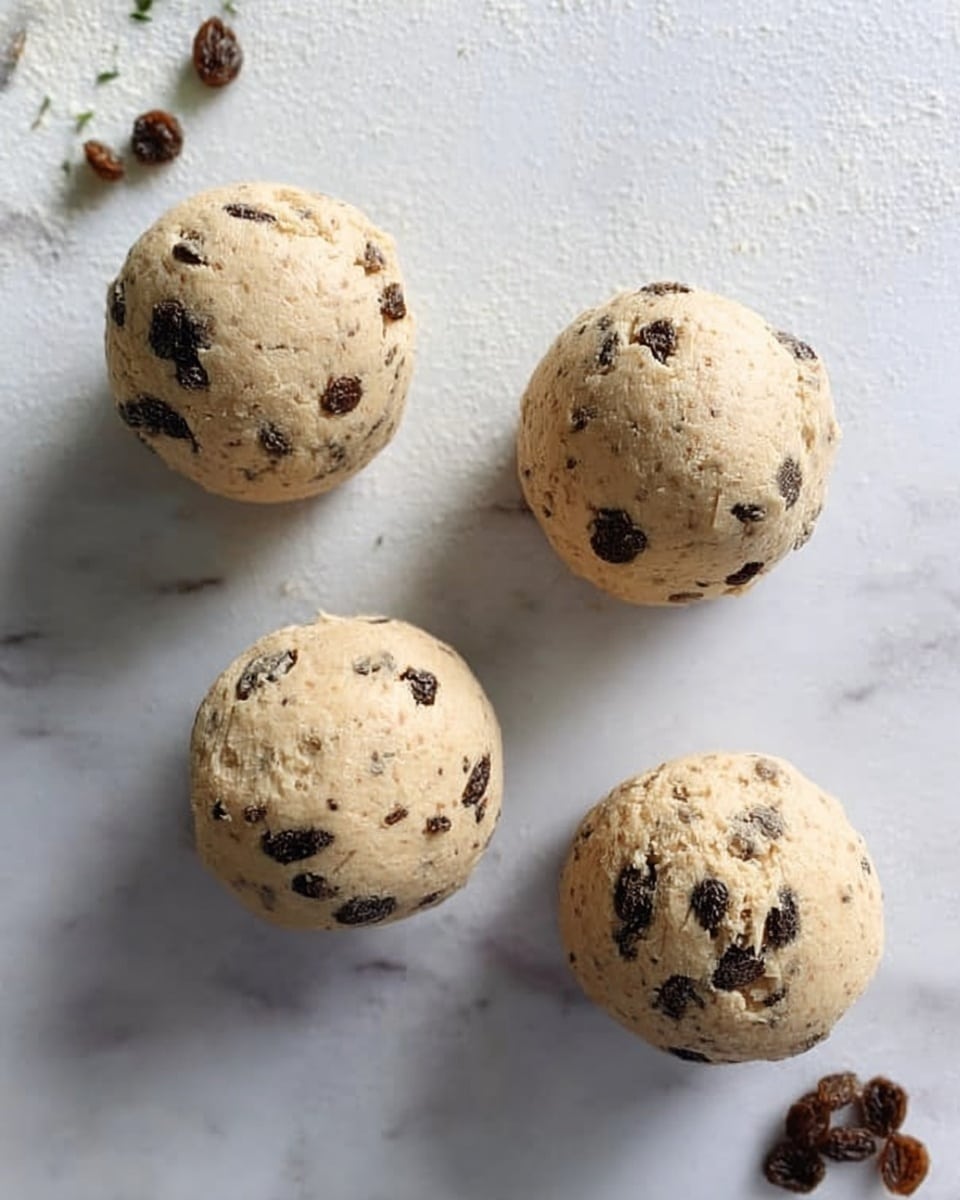 The image shows four round cookie dough balls with chocolate chips mixed inside. The dough is light beige with scattered dark chocolate spots. The balls have a rough texture and are placed spaced apart on a white marbled surface. Small crumbs and chocolate bits are scattered near the dough balls. The light is soft, making the dough look fresh and soft. photo taken with an iphone --ar 4:5 --v 7