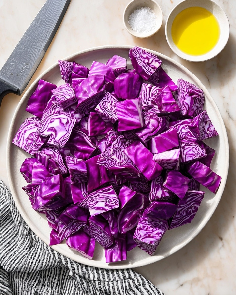 A white plate filled with many pieces of chopped purple cabbage, showing bright purple leaves with white veins, all roughly square-shaped and layered unevenly. The plate is placed on a white marbled surface with a large silver knife with a black handle on the left top side. Near the top right of the plate is a small round white bowl filled with yellow olive oil, next to a small white bowl containing white salt. A black and white striped cloth is partly visible at the bottom left corner. The light is bright and natural, highlighting the rich purple color of the cabbage and the shiny surface of the oil. photo taken with an iphone --ar 4:5 --v 7