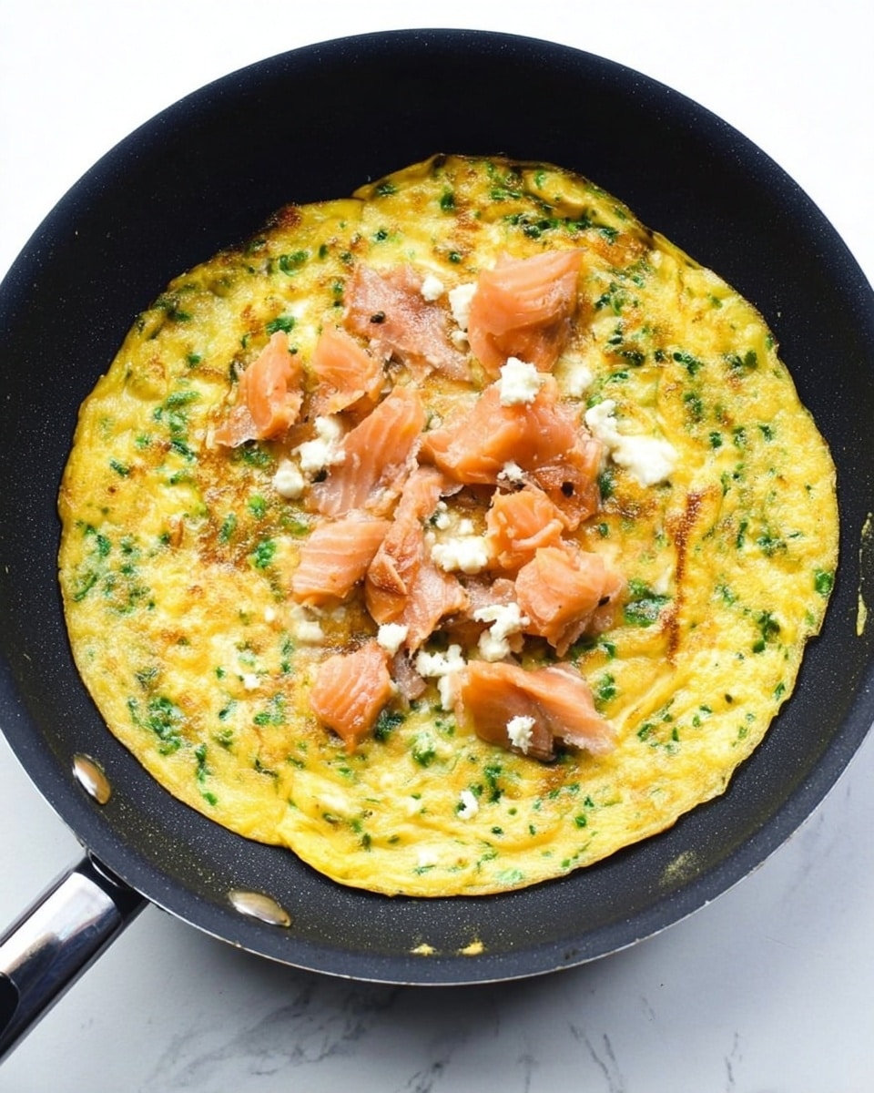 A black frying pan on a white marbled surface holds a cooked yellow egg omelet with green herbs mixed in, covering the pan evenly. On top of the omelet are uneven chunks of light pink smoked salmon and white soft cheese pieces scattered in the center. The pan handle points diagonally out of the frame. Photo taken with an iphone --ar 4:5 --v 7