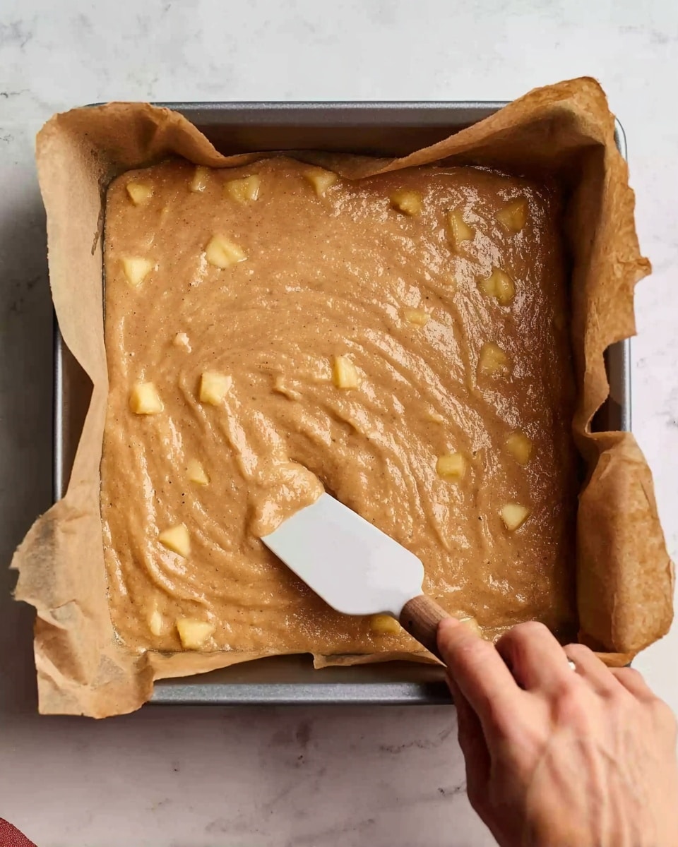 A square baking pan lined with brown parchment paper holds a thick light brown batter with small yellow fruit pieces mixed inside, the batter spread unevenly. A woman's hand is holding a white-handled spatula, smoothing the batter in the pan. The background is a white marbled surface. photo taken with an iphone --ar 4:5 --v 7